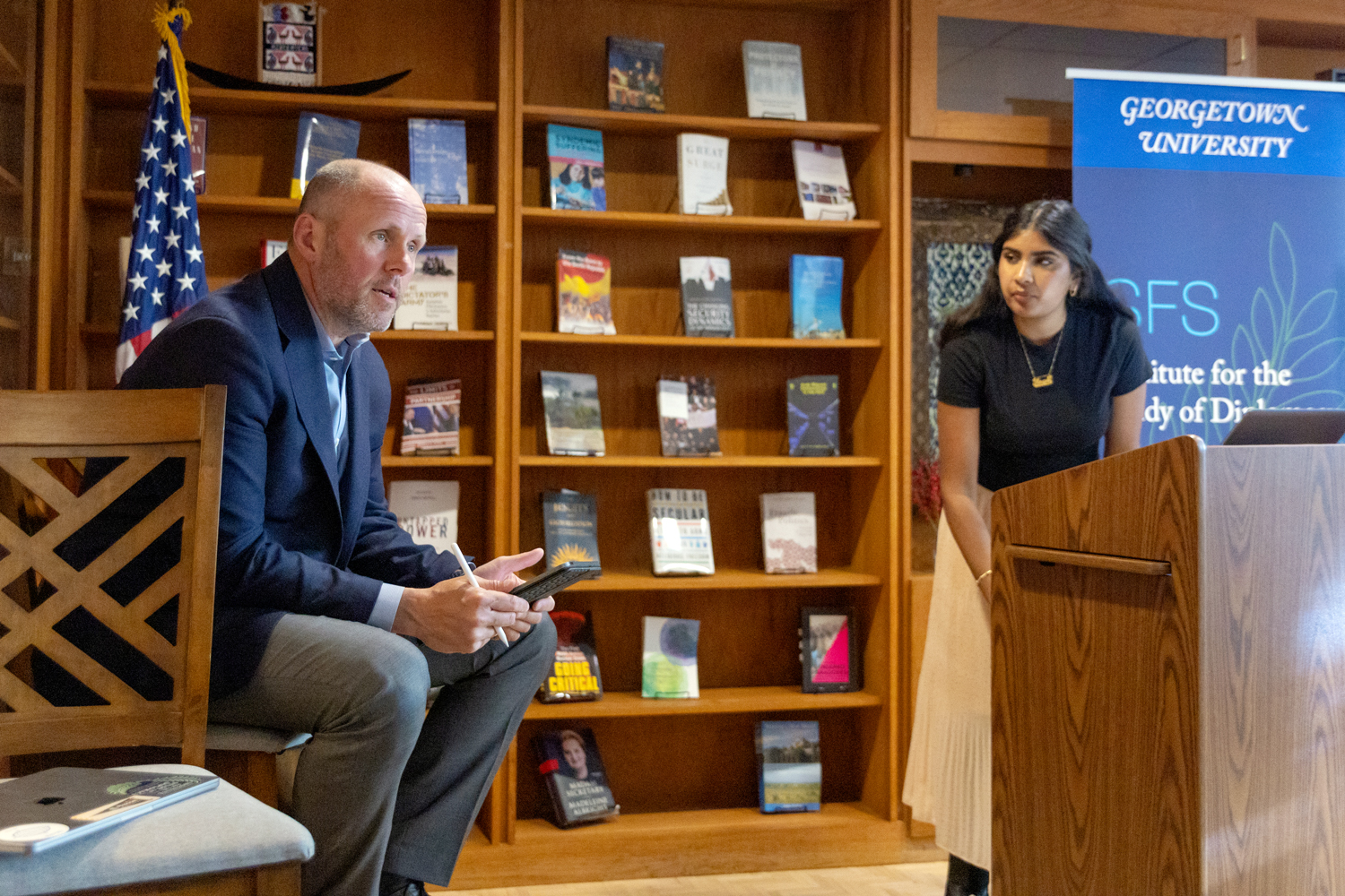 Faculty member sitting on a chair in McGhee library talking while a student in a podium listens